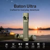 Olight Baton 4 Pro khaki flashlight on wooden surface for camping, with trailer and tent in background.