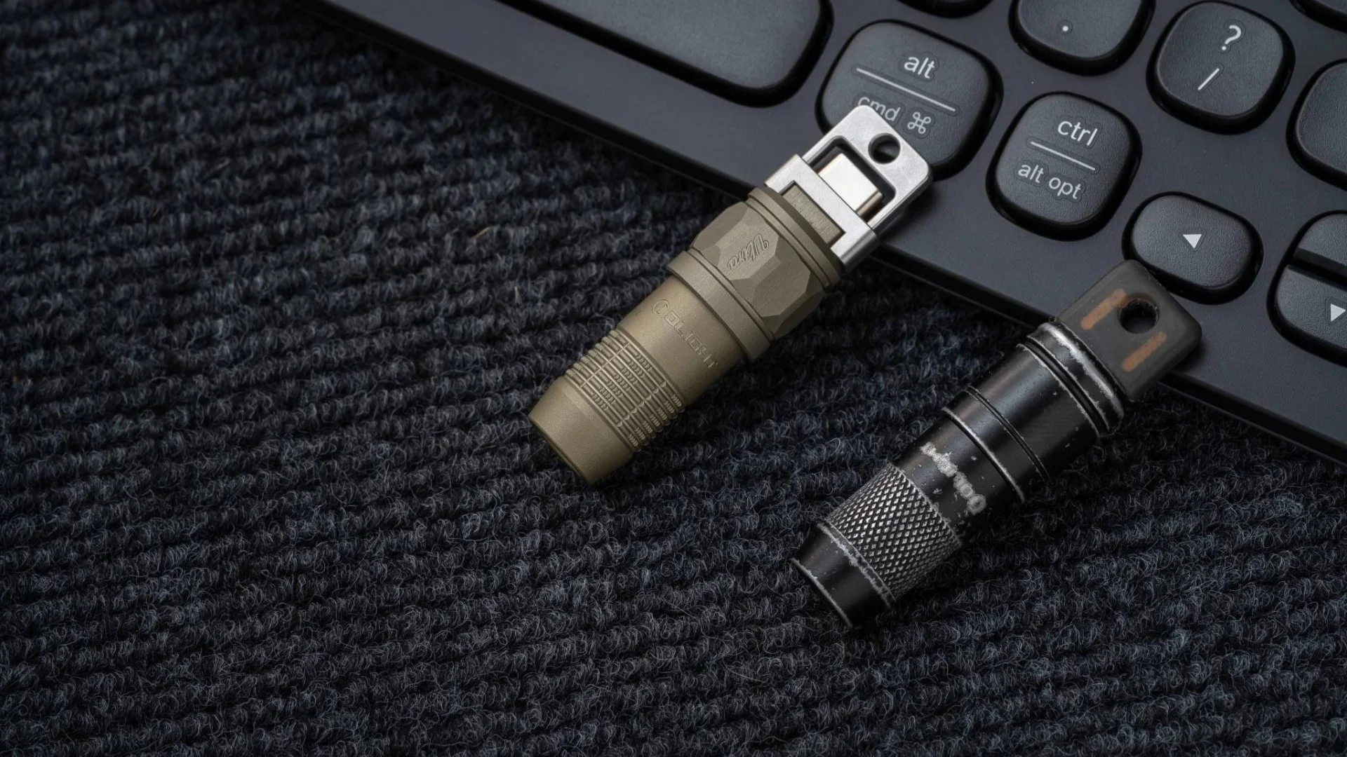 Two compact Olight flashlights, one olive green USB-C and one black, on a desk mat with a keyboard.